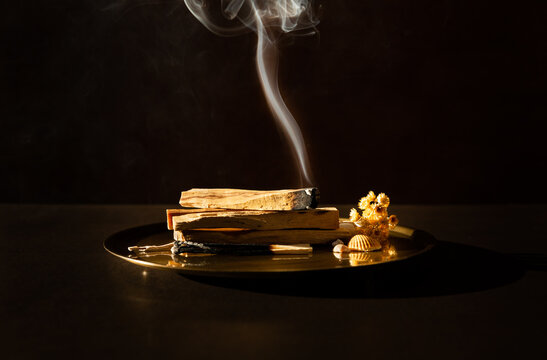 B&acirc;tons de Palo Santo sur un plateau en bronze, un b&acirc;ton en bois br&ucirc;lant et qui lib&egrave;re une fum&eacute;e blanche verticale, ambiance calme et douce, fond brun fonc&eacute;