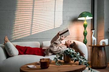 Woman lying on a cozy sofa, enjoying a peaceful moment while reading a hardback book in her sunlit living room