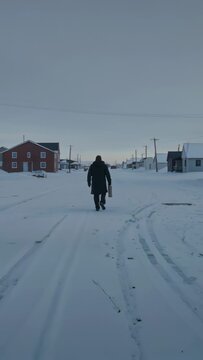 Vertical video: Starting centered walking man with case down snow road to houses in coat and boots
