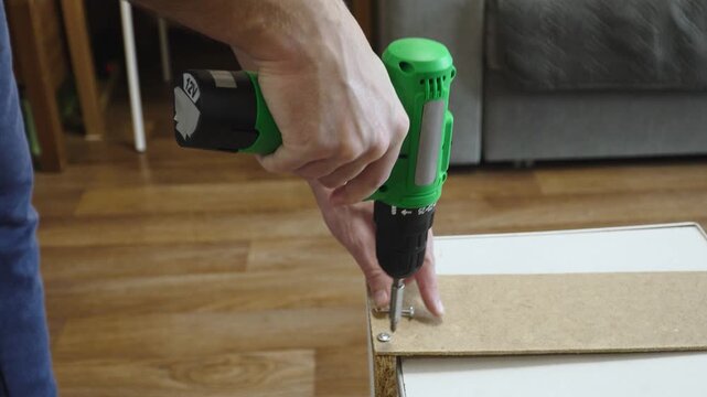 Hands of caucasian man screwing screws with screwdriver into particleboard closeup. Connecting two furniture parts and process of driving fasteners with cordless battery powered tool.