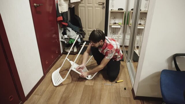 Caucasian man tightens nut on chair with back during self assembly of furniture in corridor. Wide shot of household chores with wrench in authentic house.