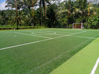 Fototapeta premium Wide angle view of an outdoor futsal court with synthetic green turf, white boundary lines, and a goal post surrounded by tropical trees.