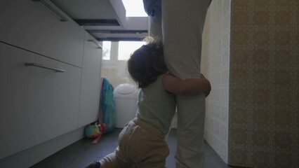 Toddler clinging to parent&rsquo;s leg in kitchen with arms raised and complaining expression, asking to be picked up while parent stands holding phone nearby