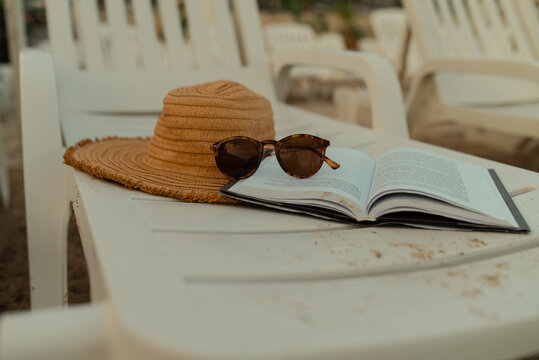 Open book, straw hat and sunglasses on a white beach chair. Summer vacation, slow living and seaside relaxation concept.