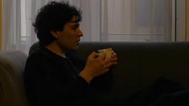 Straightening man lifting head on couch by window, cupping mug while in thought, in dark sweatshirt