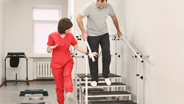 Physical therapy rehabilitation session. Female physiotherapist assisting male patient walking downstairs on training steps with handrails. Mobility recovery, balance support, gait training, medical c