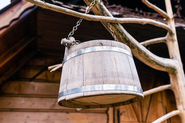 Hanging Wooden Well Bucket with Metal Bands and Chain in Front of Rustic Timber House, Vintage Rural Decoration Detail