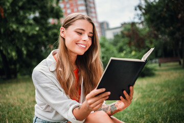 Fototapeta premium Young woman reading a notebook outdoors in a sunny park smiling and enjoying a relaxed moment in the city park setting