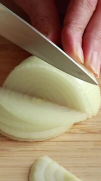 Close-up of woman&rsquo;s hands slicing a raw peeled yellow onion into half-circle slices on a wooden cutting board