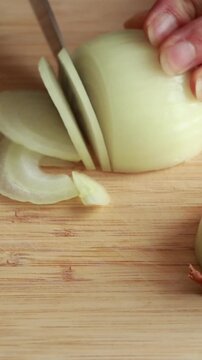 Close-up of woman&rsquo;s hands slicing a raw peeled yellow onion into half-circle slices on a wooden cutting board