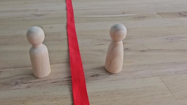 Two wooden figures standing on opposite sides of a red ribbon on a wooden surface representing division and separation