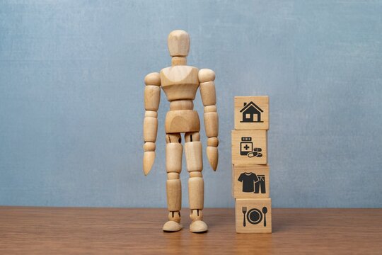 A wooden doll stands next to stacked blocks with icons for housing, healthcare, clothing, and food. Basic human needs, cost of living, and life essentials concept.