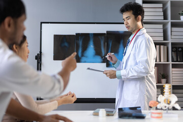 Obraz premium Asian doctor showing radiography to coworker and explaining diagnosis, holding clipboard and pointing at whiteboard with pen during meeting in hospital office.