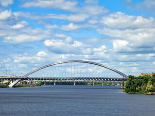 Obraz premium A bridge arch over river. An openwork architectural engineering structure for logistics cargo transportation across body of water. Lower angle shot shore, perspective road. Cumulus clouds landscape