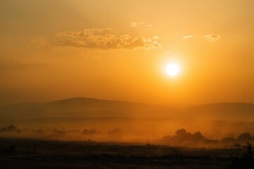 Obraz premium Hazy sunset panorama: warm orange tones, smoky clouds crossing the sun, serene distant hills