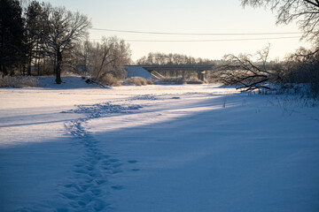 snow covered river, frozen water surface covered with snow, bright winter morning, tree shadows on snow, transport bridge ahead, footprints in snow