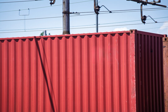 Close-up of a red corrugated shipping container on a freight train, with overhead lines and rail yard elements, illustrating cargo transport, imports, exports and logistics.