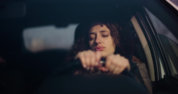 A young woman with visible braces looks sleepy as she drives her car during the evening. The reflection of the sunset can be seen in the window.