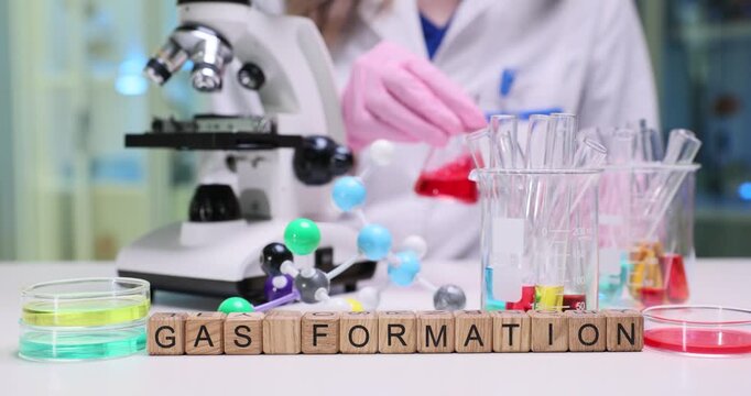 Wooden cubes spell phrase Gas Formation on lab desk with glassware. Scientist in protective gloves studies bubbling chemical reaction under microscope