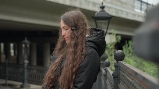 young woman walking on urban bridge, contemplative portrait sequence showing traveler pausing by railing under overpass, hooded coat and long hair, soft morning light, muted tones, distant lamp