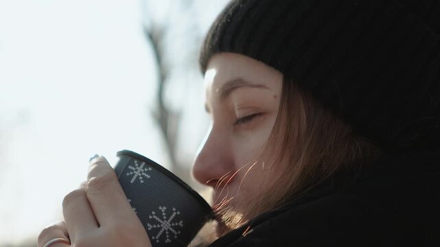 A beautiful young woman drinking hot coffee in a winter park outdoors. A walk outdoors on a cold, frosty day.