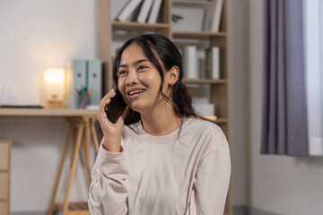 Fototapeta premium Cheerful young asian woman talking on mobile phone having conversation with happy smile expression while relaxing at cozy home living room during leisure time