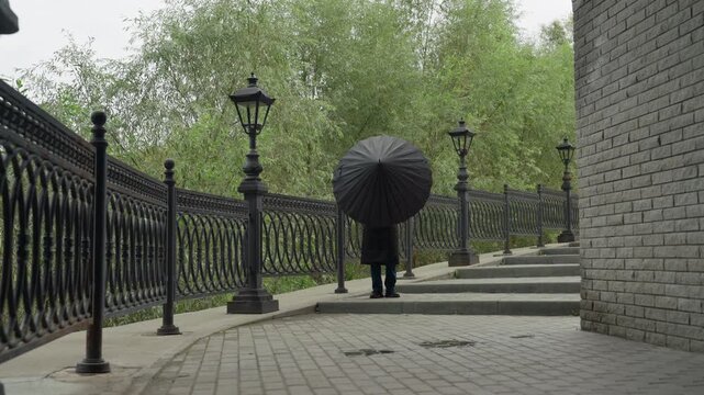 woman walking on iron bridge umbrella, black coat, vintage lamps, cobblestone path, overcast sky, light drizzle, solitary reflective stroll, slow cinematic pan to closeup umbrella spokes, melancholic