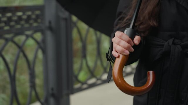 Closeup grip on wooden umbrella handle, tailored black coat, decorative balcony railing, subtle movement suggesting walking or waiting, urban morning light, cinematic color, soft bokeh, narrative