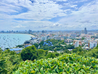 High angle panoramic view of Pattaya City and crescent moon bay with many boats in the ocean. The image showcases the dense cityscape meeting the blue sea under a cloudy sky. Captured in Pattaya © Bachhuyhoang