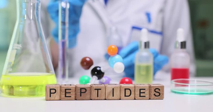 Wooden cubes spell word Peptides on laboratory table. Scientist in blue scrubs works with molecular model and colorful solutions during research