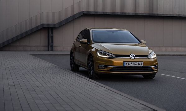 amber VW Golf in front of beige wall. Front three quarter view of Volkswagen hatchback in urban setting - parked near a curb and beige-colored wall. Kyiv, Ukraine - June 07 2025