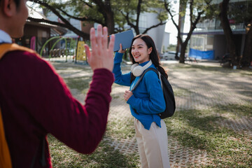 Naklejka premium Happy young Asian student carrying a backpack and tablet, smiling and waving to a friend on a university campus
