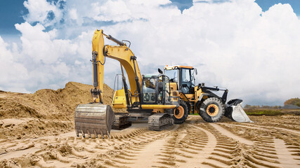 Heavy machinery is at work on sandy ground, leaving deep tracks. A large pile of sand is visible in the background indicating ongoing construction activities