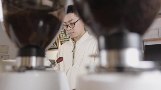 Focused young asian barista meticulously leveling ground coffee in portafilter, preparing to brew a perfect espresso with professional equipment in a contemporary coffee shop