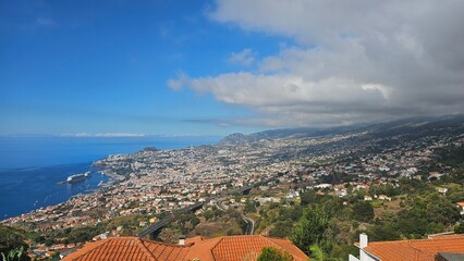 Wide daytime panorama of Funchal, Madeira, showing the Atlantic Ocean harbor, hillside neighborhoods, winding roads, and lush green slopes under a bright blue sky with scattered clouds. © Sebastiaan89
