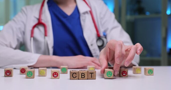 Wooden cubes spell abbreviation CBT on white medical desk. Female doctor moves emoji cubes symbolizing mental health session for patient in clinic
