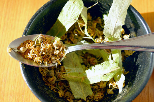 Spoon holding dried linden blossoms above a bowl in strong sunlight. Illustrates modern seasonal home remedy, gentle self-medication alternative, calming herbal infusion, and domestic health practice.