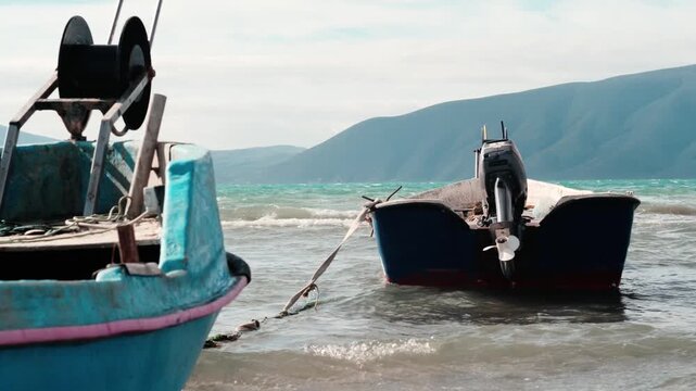 Two small fishing boats floating in shallow sea water near the shore, with visible outboard motor, ropes and metal winches. Natural daylight, layered depth, calm waves and balanced coastal composition