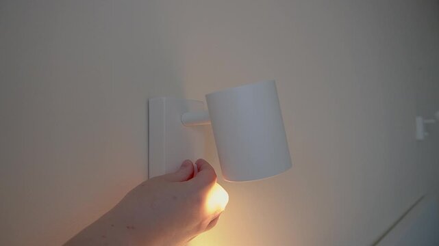 The hand turns on the wall lamp. White wall lamp with a rotating shade, warm glow, light wall, close-up.