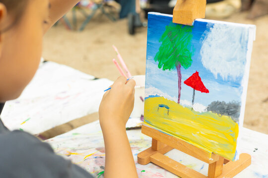 Close-up and selective focus of young girl painting a palm tree and sea with beautiful color on canvas. This shows creativity of a child's artistic hobby in a natural, outdoor beach setting.