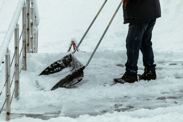 Person uses a wide snow shovel to remove fresh snow from a path, suggesting routine municipal or property upkeep after snowfall. The scene communicates cold-weather labor, preparedness, and preventing