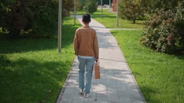 asian male illustrator walking away down pathway in sunlit park, carrying leather portfolio briefcase, casual jacket, retreating from camera with quiet reflective mood, lush greenery and soft shadows