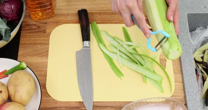 Hands use peeler to slice fresh squash into thin ribbons for cooking at home. Cutting board holds peeled strips of zucchini by large knife time-lapse