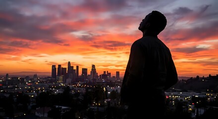 Fototapeta premium Silhouette of contemplative man overlooking illuminated city skyline during dramatic orange and pink sunset with urban lights twinkling below.