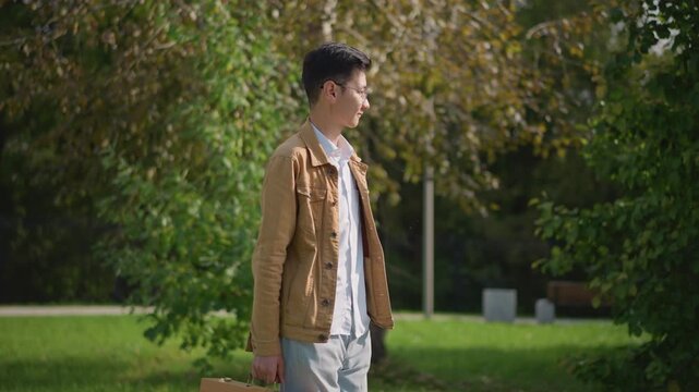 young asian man scanning park foliage, wearing brown jacket and glasses, walking past pavilion and shrubs, glances toward distant pathway, casual pace, ambient sunlight and soft shadows, background