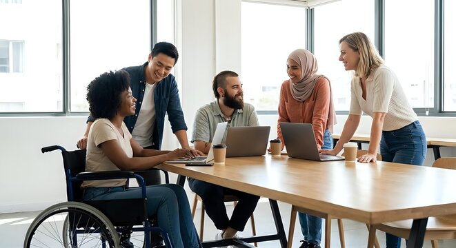 Diverse business team collaborating around laptop in modern office with natural light. Includes woman in wheelchair promoting workplace inclusion and accessibility.