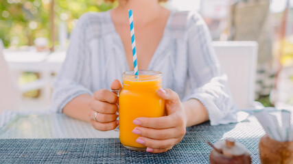 Woman holding orange juice in mason jar at sunny cafe terrace, healthy summer drink concept.
