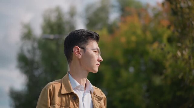 asian man on sunlit street, contemplative portrait. young asian artist wearing glasses and tan jacket gazes across neighborhood with soft bokeh and leafy backdrop, warm cinematic tones, casual