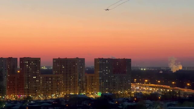 Vibrant orange and blue sunset over a modern city skyline with illuminated residential apartment buildings and a lone airplane in the sky, capturing an atmospheric urban evening.
