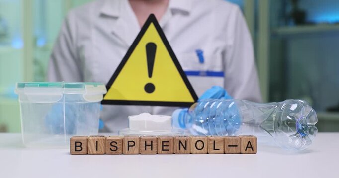 Wooden blocks spell word Bisphenol-a by plastic bottle and international warning sign. Lab worker positions warning sign checking plastic bottle label